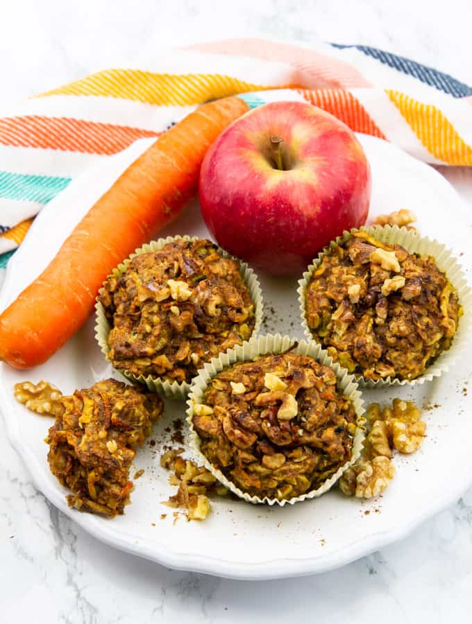 three zucchini carrot muffins on a white plate with an apple and a carrot on a marble countertop 