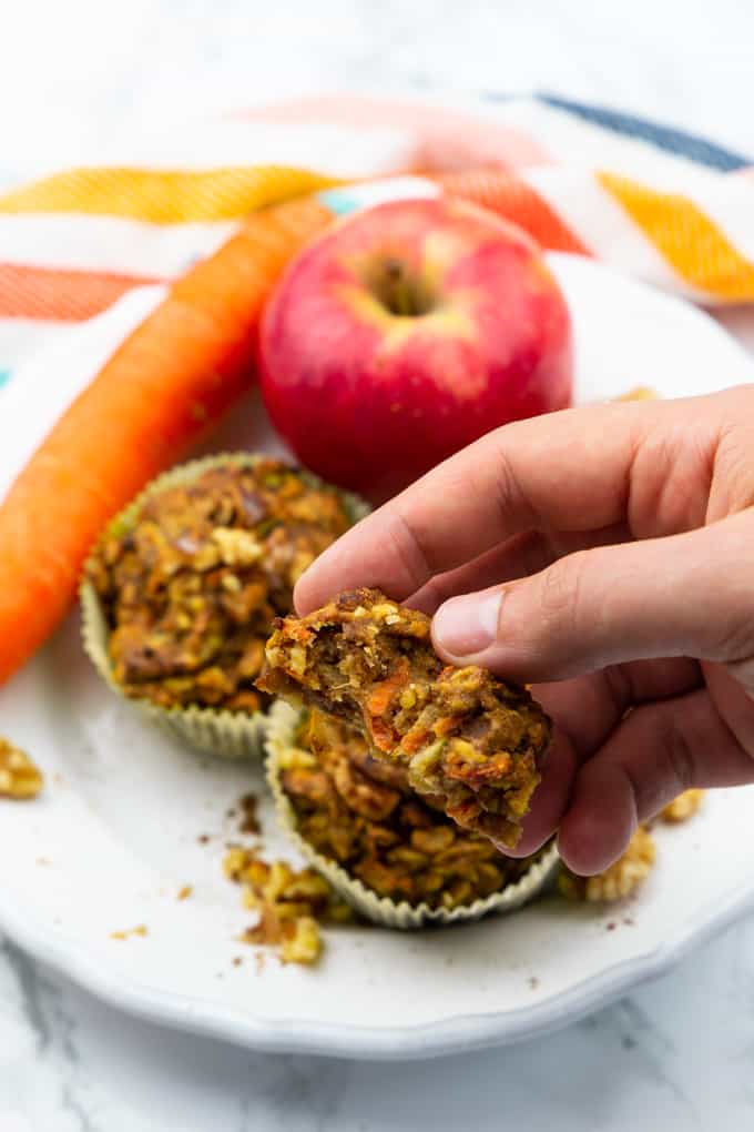 a hand holding half a zucchini carrot muffins with a plate with more muffins, an apple, and a carrot in the background