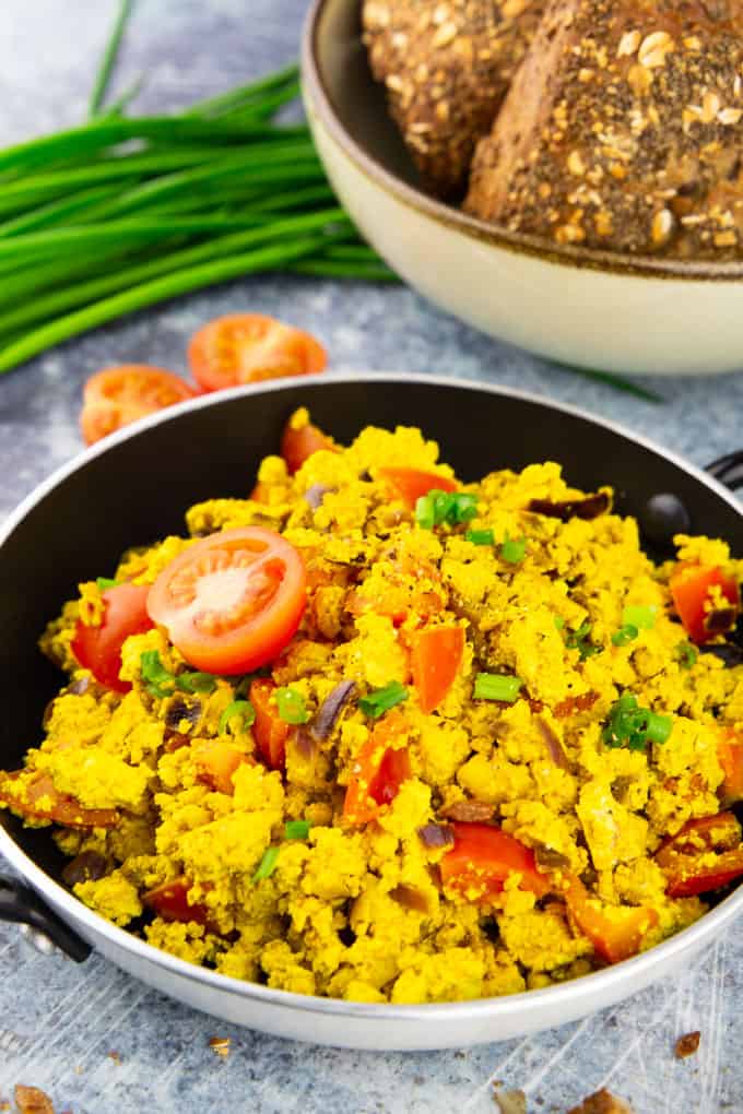 tofu scramble with tomatoes in a small pan on a blue counter top with a bowl with buns and fresh chives in the background