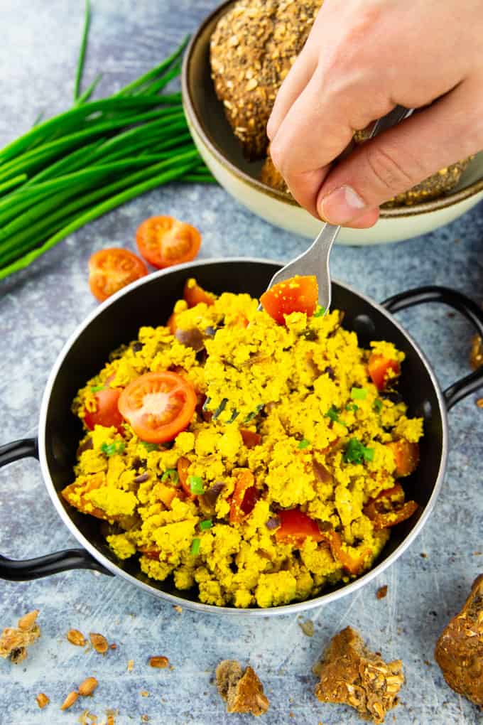 a handing holding a fork in a small pan with tofu scramble on a blue counter top with buns and fresh chives on the side 