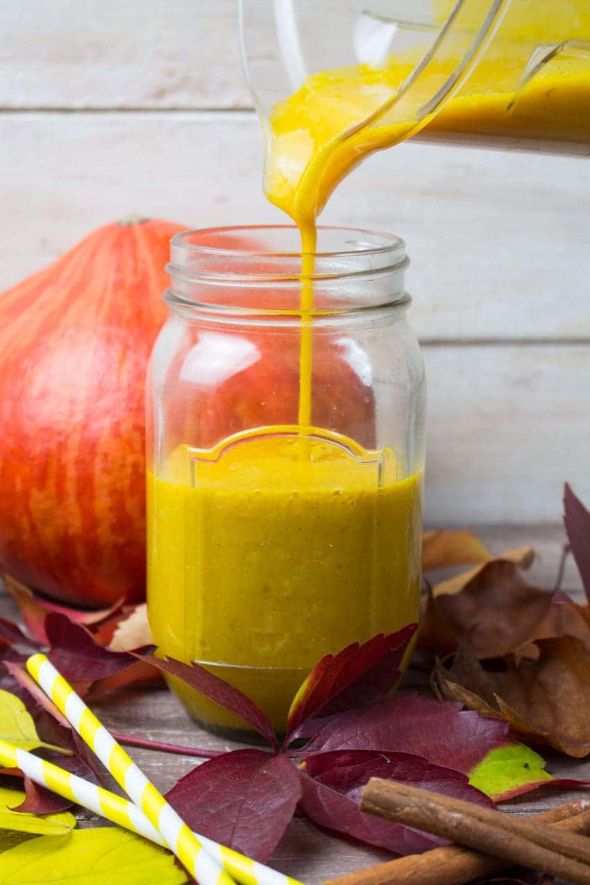 pumpkin smoothie is being poured from a Vitamix container into a jar with red leaves and a pumpkin in the background