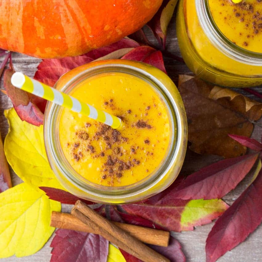 two glasses of pumpkin pie smoothie with a yellow and white straw on red and yellow fall leaves with two cinnamon sticks on the side 