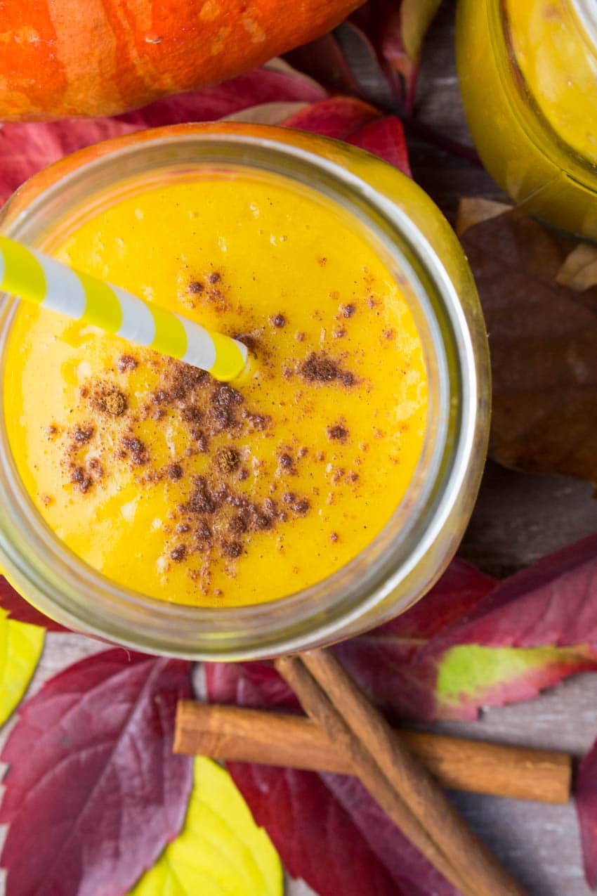 a glass of pumpkin smoothie with a yellow straw and cinnamon on top on red leaves with a pumpkin in the background
