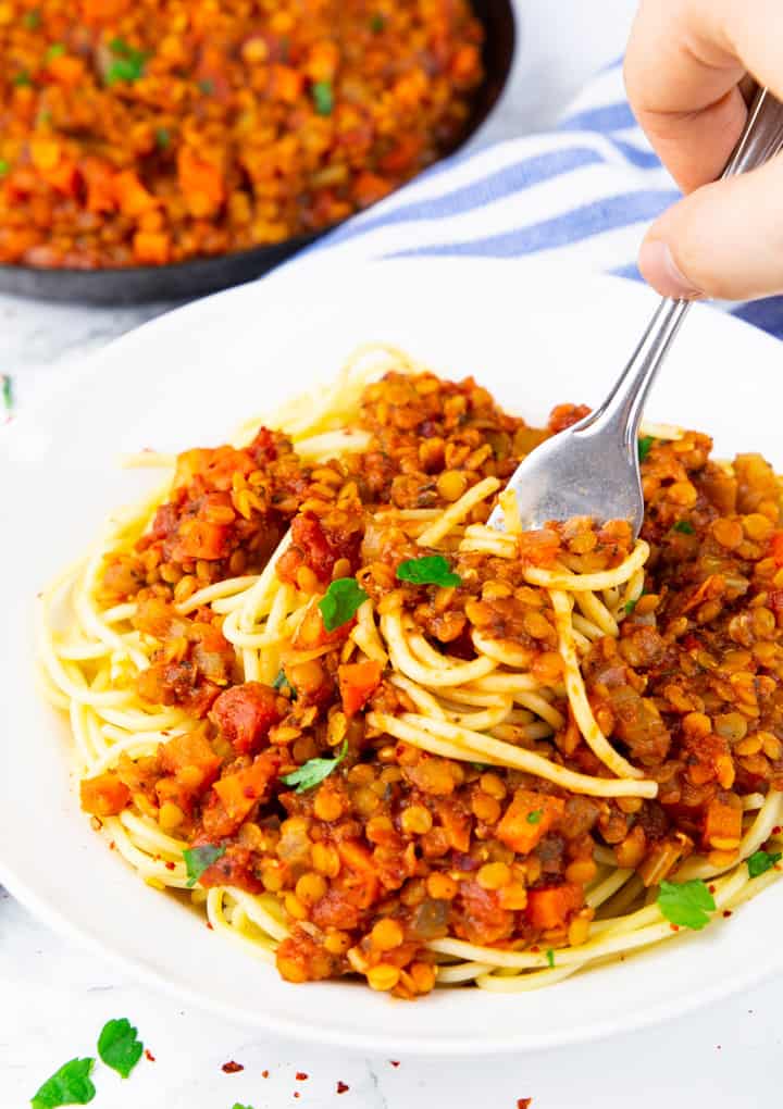 a white bowl of lentil bolognese with spaghetti with a hand holding a fork and a pan of bolognese sauce in the background