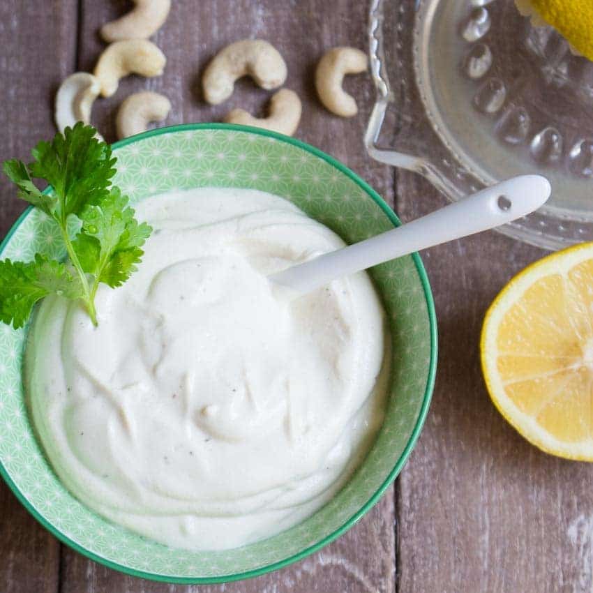 vegan sour cream in a green bowl with a white spoon on a wooden counter top with cashews and a lemon half on the side