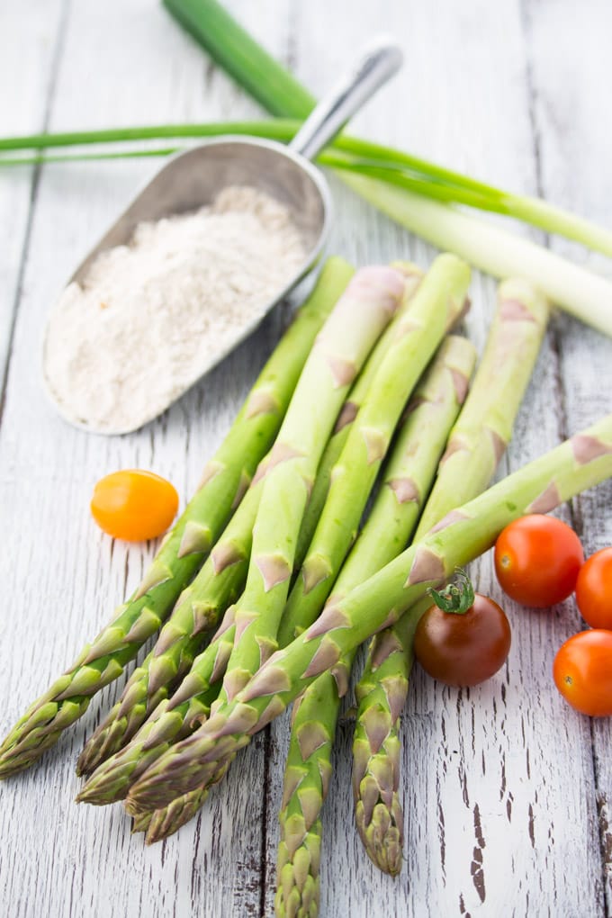 a bunch of green asparagus on a white wooden countertop with flour, green onions, and cherry tomatoes on the side and in the background