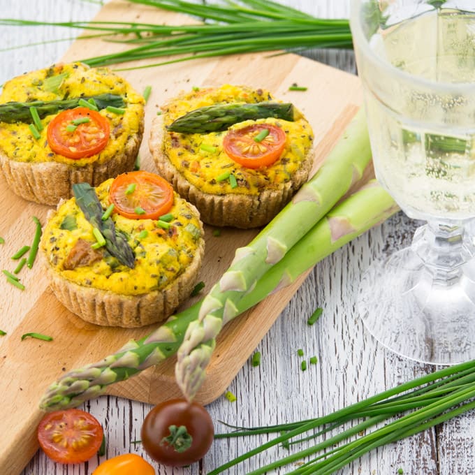 Three Vegan Quiche with asparagus on a wooden board with green asparagus, chives, tomatoes, and a glass of white wine on the side