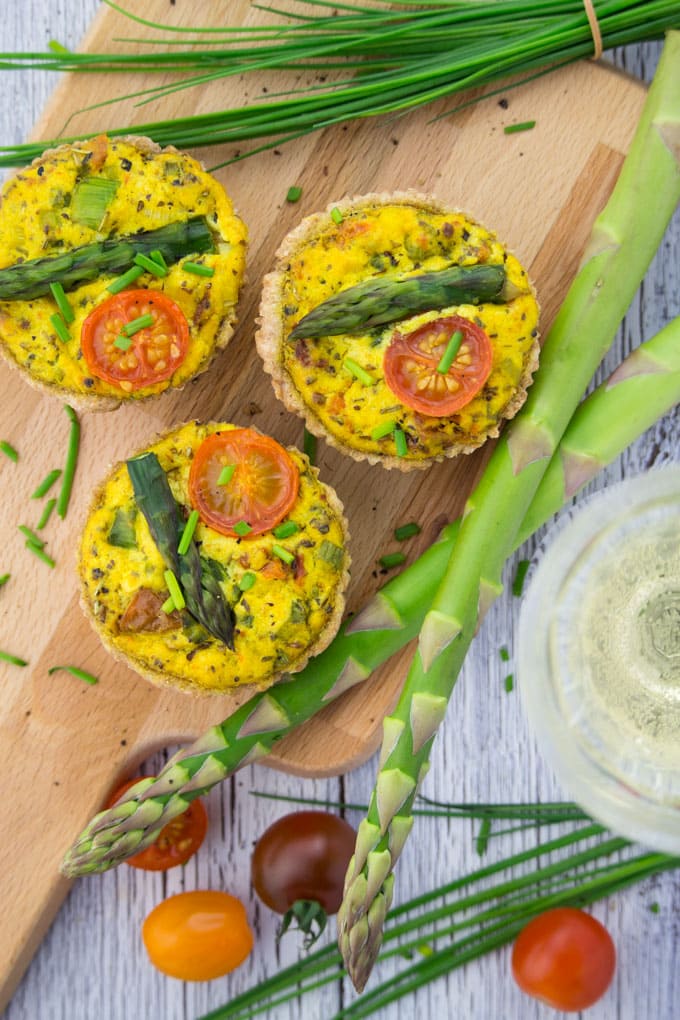 Three Vegan Quiche with asparagus on a wooden board with green asparagus, chives, tomatoes, and a glass of white wine on the side