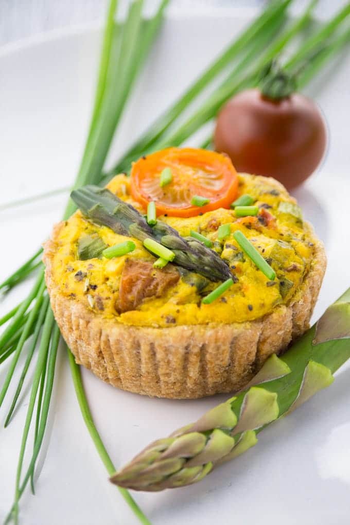 a vegan mini quiche on a white plate with chives, green asparagus, and a cherry tomato on the side 