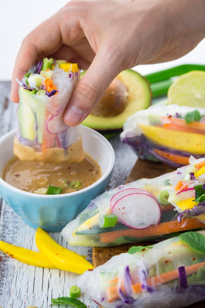 a hand dipping a rice paper roll into a small bowl with peanut sauce with more rice paper rolls in the background