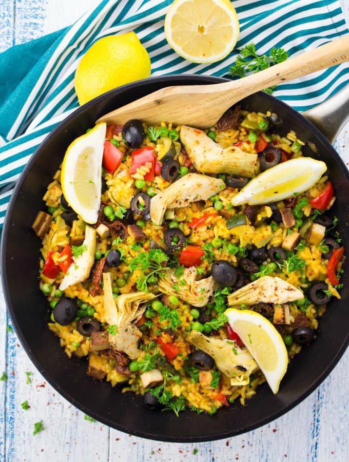 Vegan Paella in a black pan with a wooden spoon on a white and blue wooden countertop with two lemons in the background