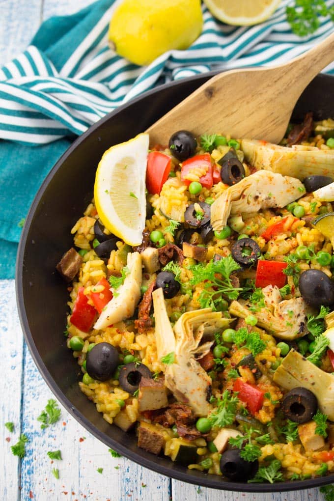 Vegan Paella in a black pan with a wooden spoon on a white and blue wooden countertop