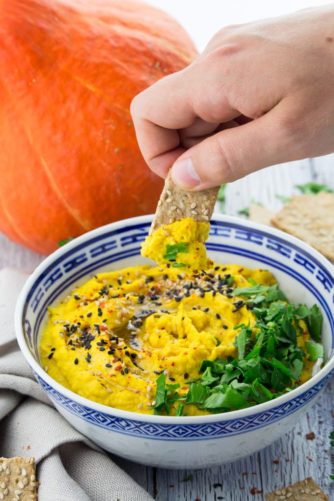 a hand dipping a sesame cracker into a bowl of pumpkin hummus with a pumpkin in the background