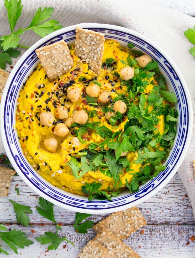 pumpkin hummus in a blue and white bowl with fresh parsley, sesame seeds, and sesame crackers on top
