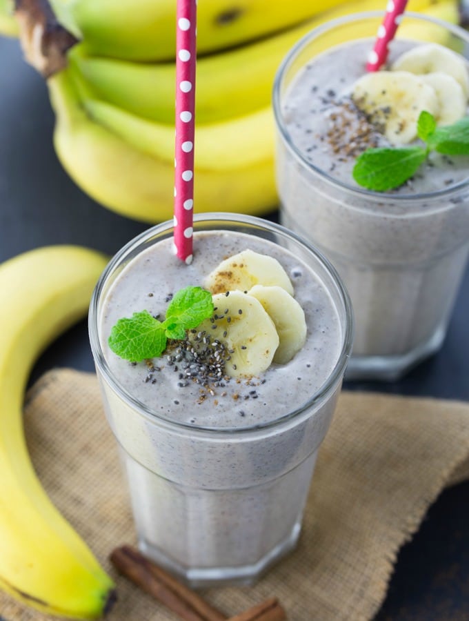 Two glasses with banana smoothie on a black counter top with bananas in the background