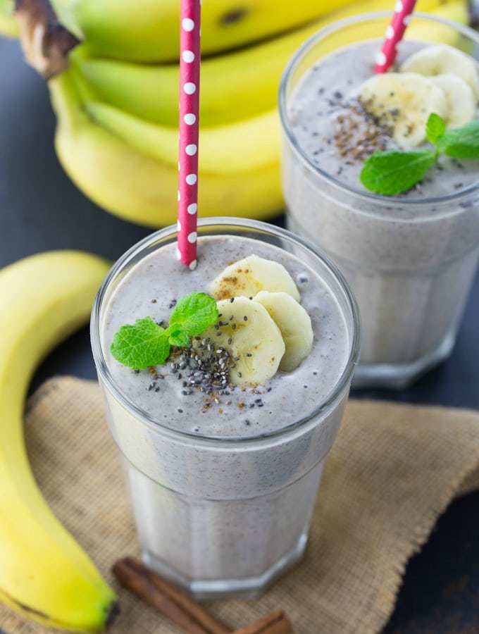 Twee glazen met bananensmoothie op een zwart aanrechtblad met bananen op de achtergrond