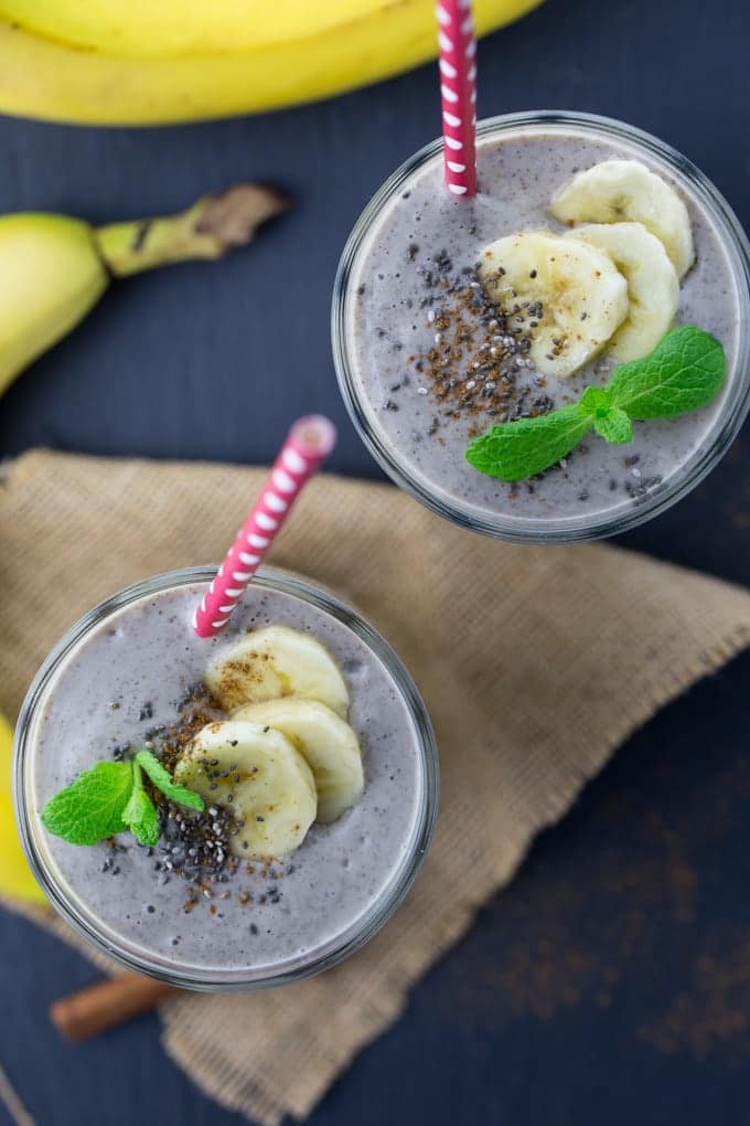 Two glasses of vegan banana smoothie on a black counter top with a brown napkin 