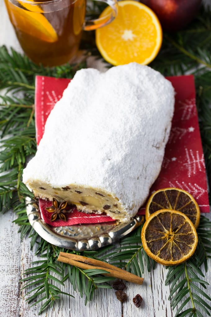 a German stollen on a plate with a red Christmas napkin underneath and evergreens, dried orange slices, and cinnamon sticks on the side