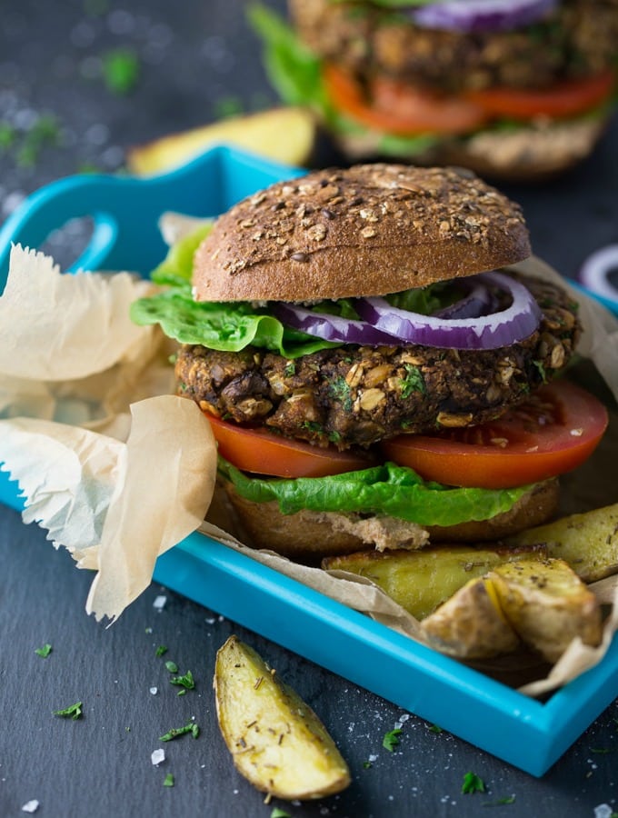 een champignonburger met tomaten, sla en rode ui op een blauwe serveerschaal met perkamentpapier en aardappelpartjes aan de zijkant