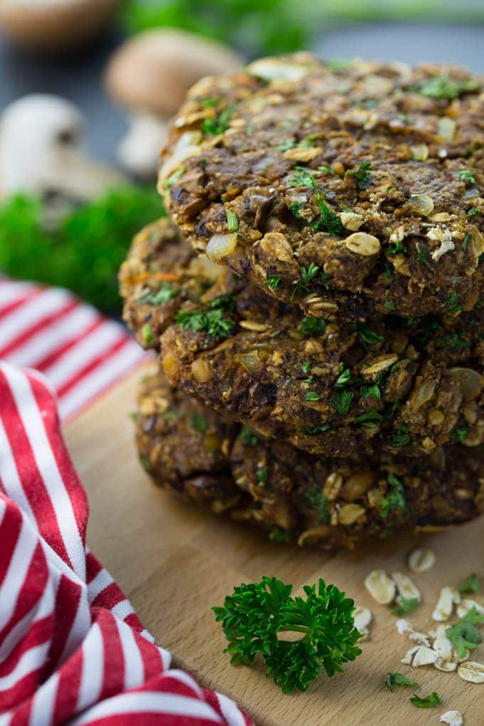 uma pilha de três hambúrgueres de cogumelos numa tábua de madeira com cogumelos e salsa ao fundo