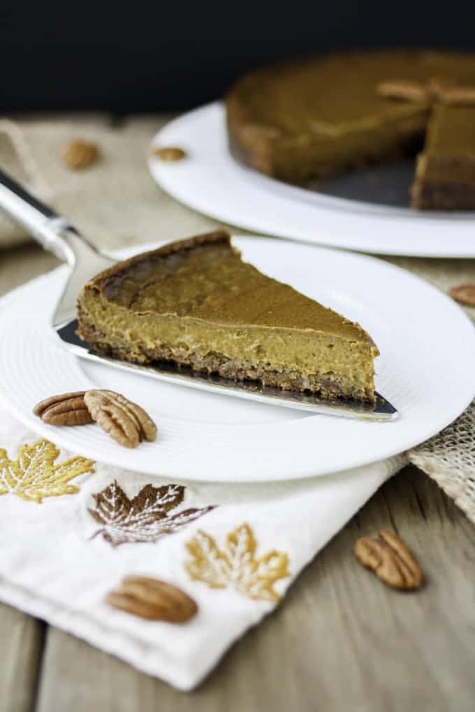 a piece of vegan pumpkin pie on a white plate on a fall-theme table cloth with more pumpkin pie in the background 