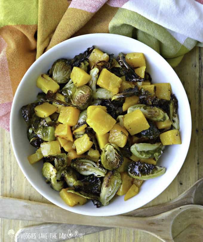 roasted Brussels sprouts and butternut squash in a white bowl with an orange and green table cloth underneath and two wooden spoons on the side