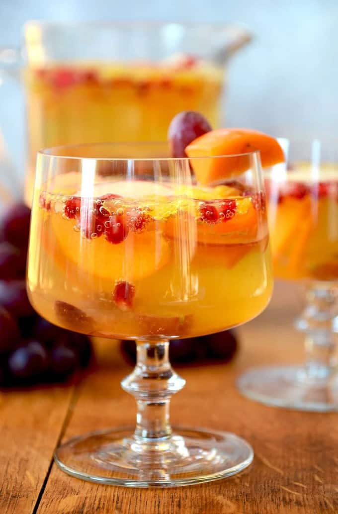 a glass of cider sangria with grapes, persimmons, and oranges on a wooden board with a pitcher of more sangria in the background