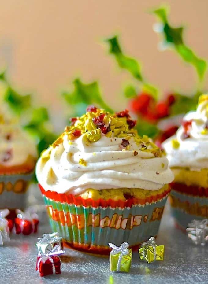 a vegan orange muffin with vanilla cream and pistachios on top with two other muffins and Christmas decoration in the background and on the side 