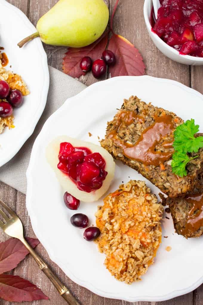 two slices of lentil loaf with vegan gravy and mashed sweet potato and cranberry sauce on a white plate on a wooden board with a fork and a small bowl of cranberry sauce on the side