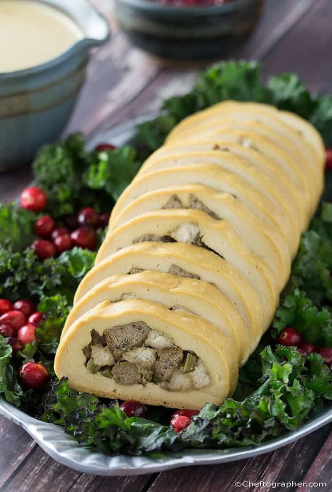 a vegan turkey roll on a bed of kale and fresh cranberries on a serving plate with a bowl of gravy in the background