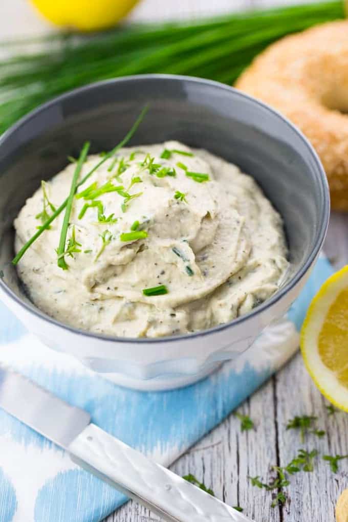 vegan cream cheese in a grey and white bowl with chives, lemons, and a bagel in the background