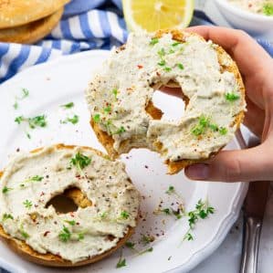 a hand holding a bagel with vegan cream cheese over a white plate
