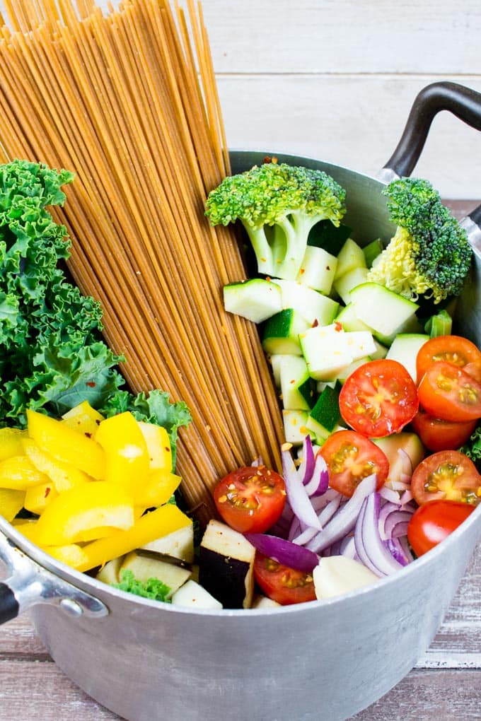 a pot with uncooked spaghetti, cherry tomatoes, broccoli, bell pepper, zucchini, and kale on a wooden board 