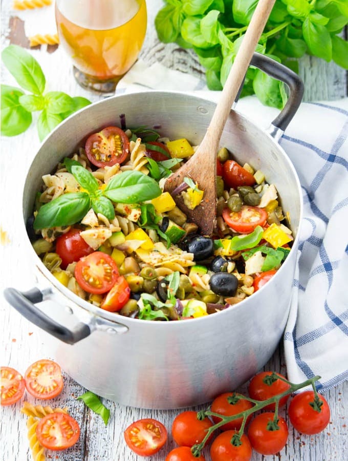 a pot of Italian pasta with olives, cherry tomatoes, bell peppers, and capers on a white wooden board with fresh basil, tomatoes, olive oil, and a blue and white dishcloth on the side 