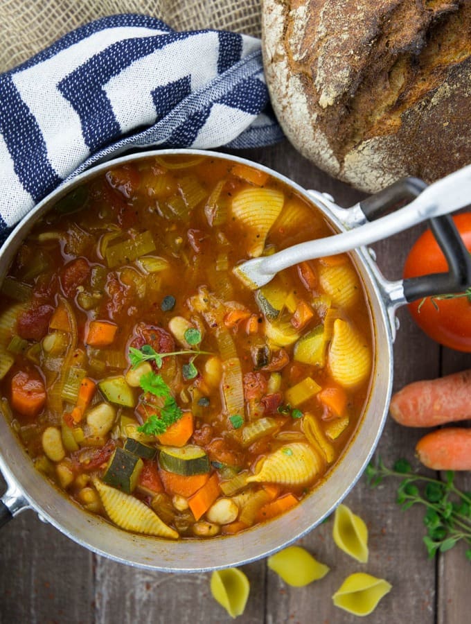 a pot with vegan minestrone soup on a wooden countertop with a loaf of bread on the side
