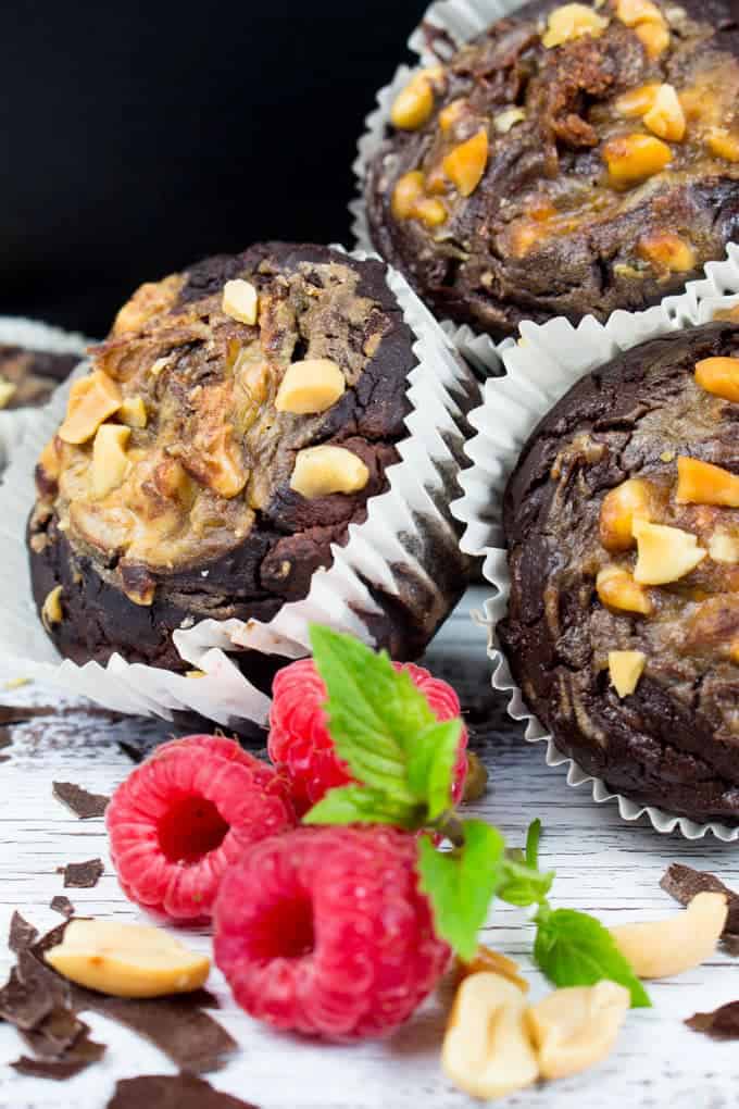 three vegan chocolate muffins with peanuts on top on a white wooden board with three raspberries in front 