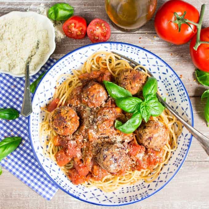 spaghetti with vegan meatballs and marinara sauce in a blue and white bowl on a wooden board with tomatoes, basil leaves, and a bowl of vegan Parmesan cheese in the background