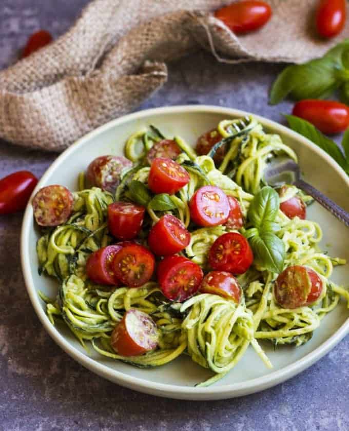 a plate with basil zoodles and cherry tomatoes on a grey counter top with basil leaves and tomatoes in the background