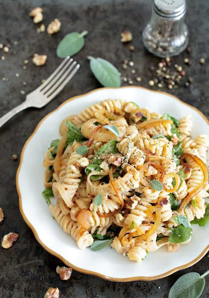 spinach and caramelized onion pasta on a white plate with a fork, fresh sage, and a pepper shaker on the side 