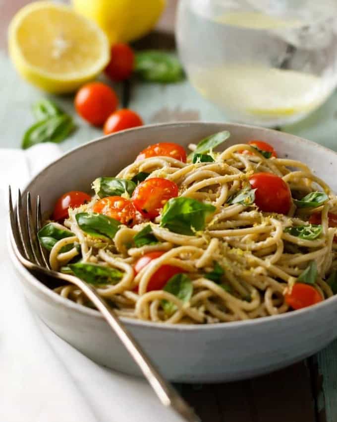 a bowl of lemon basil pasta with walnut parmesan and cherry tomatoes with a fork on the side and a glass of water and two lemons in the background