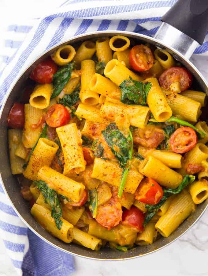 a pot of vegan one pot pasta with rigatoni, spinach, and cherry tomatoes on a marble counter top with a blue tablecloth