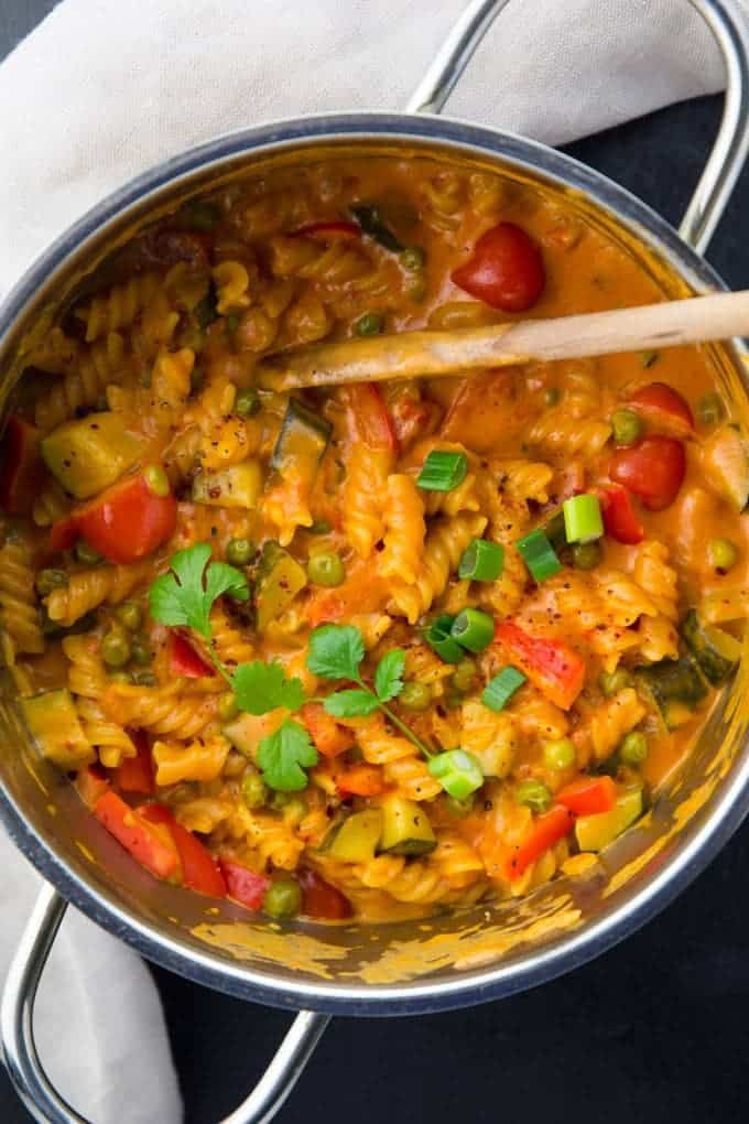 a pot of vegan one pot pasta with peas, zucchini, and bell peppers and a wooden spoon on a black counter top with a grey dish cloth