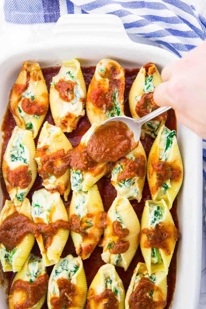 a casserole dish of vegan stuffed shells with spinach and a hand with a spoon putting tomato sauce on top of the shells 