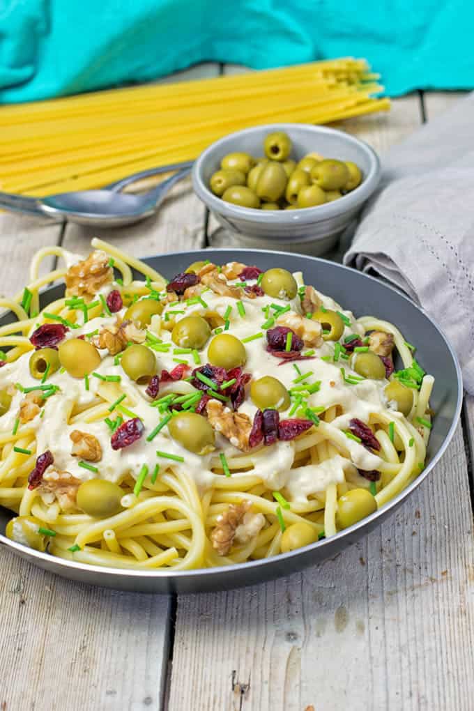 a bowl of mediteranean pasta with creamy sauce, green olives, and dried tomatoes in a grey plate with a small bowl of olives, a spoon, and uncooked spaghetti in the background 