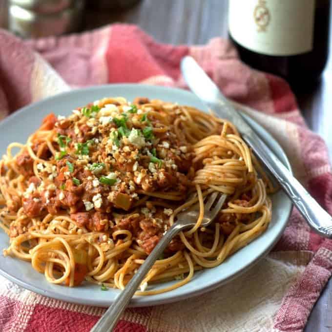 a plate of spaghetti with vegan tempeh ragu on top of a red and white tablecloth and a bottle of red wine in the background