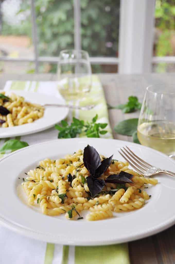 two white plates with herb gemelli pasta on a table with two glasses of wine and a window in the background