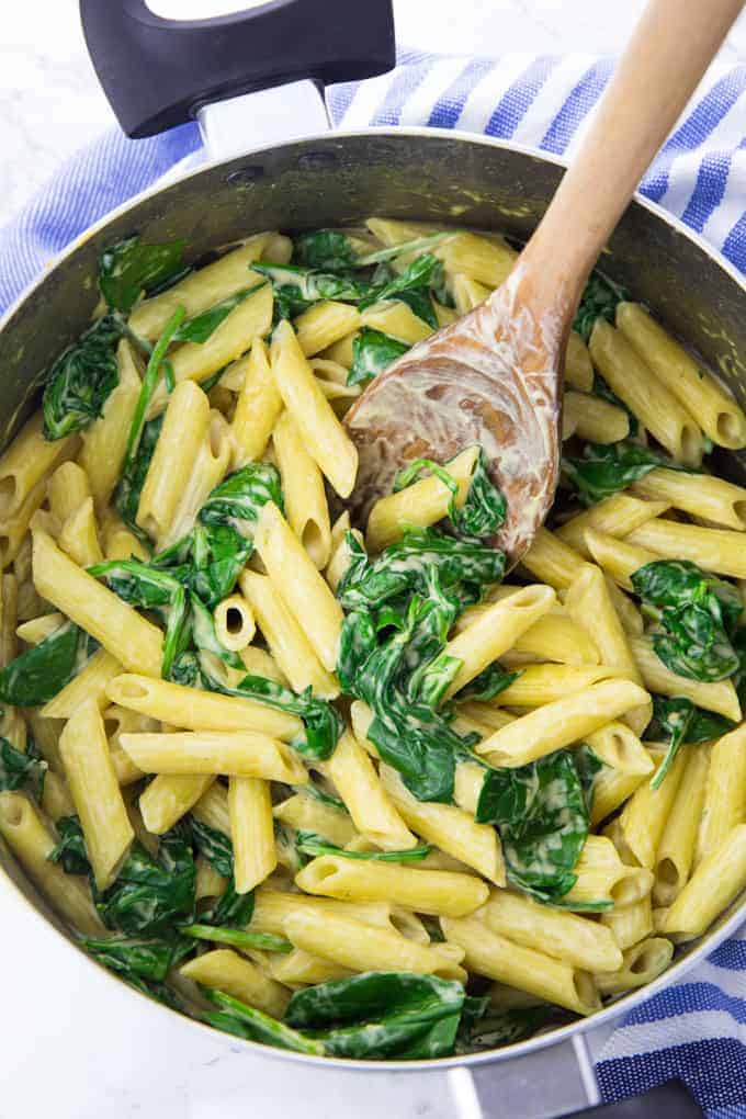 a pot of spinach artichoke pasta with a wooden spoon on a marble counter top with a blue dishcloth 