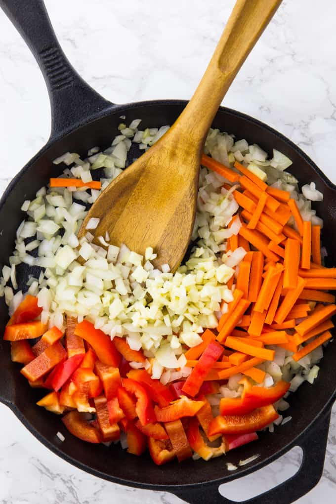 Sauted onions, garlic, and carrots and red bell pepper stripes in a cast iron skillet with a wooden spoon