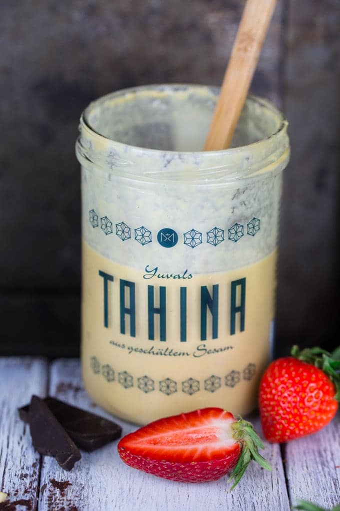 a jar of tahini with a wooden spoon on a white wooden countertop with dark chocolate and two strawberries on the side 