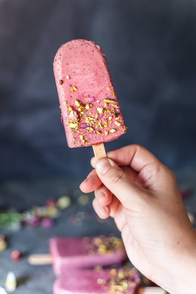 a hand holding a wild strawberry popsicles in front of a dark grey background with two more popsicles in the background 